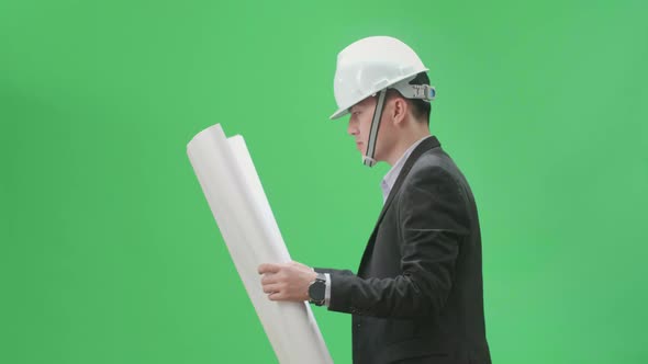 Asian Chief Engineer In The Hard Hat Holding And Looking At Blueprint While Walks On Green Screen