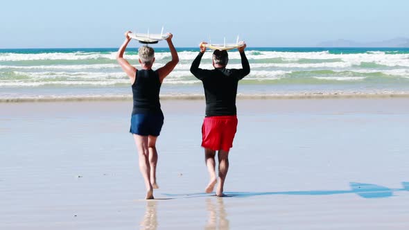 Senior couple carrying surfboard over head while running towards the sea