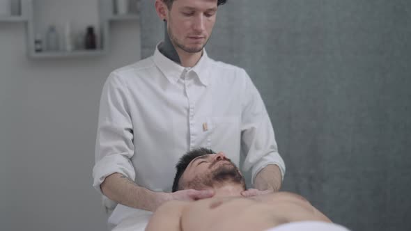 Handsome Caucasian Man Lying on Massage Bed As Concentrated Male Masseur Massaging Neck Muscles in