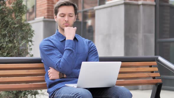 Pensive Young Man Working on Laptop Sitting Outdoor on Bench