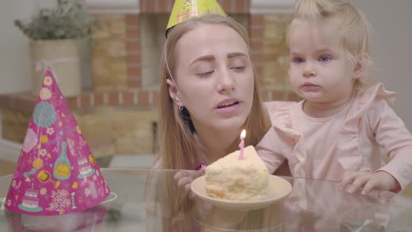 Close-up of Young Caucasian Woman in Party Hat and Little Baby Girl Blowing Candle on the Piece
