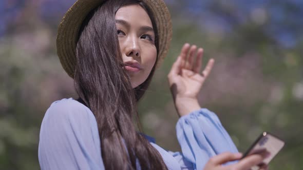 Young Asian Woman in Straw Hat Texting on Smartphone Smiling Standing in Sunlight in Spring Sakura