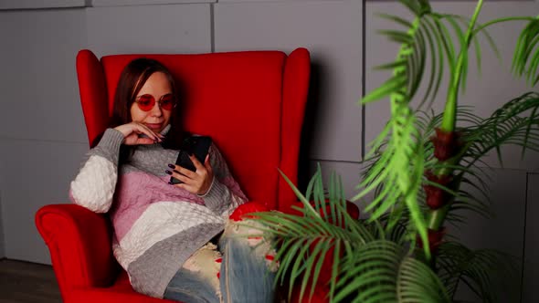 Woman in stylish sunglasses sitting in armchair with smartphone.