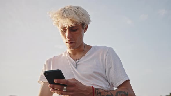 Portrait of Young Fashionable Blonde with Tattoos Smokes a Cigarette and Holds a Smartphone in His