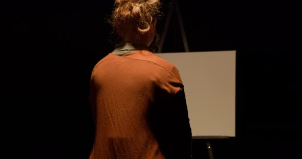 Depressed woman wearing orange sweater in scary dark black void in front of an easel, camera pushes