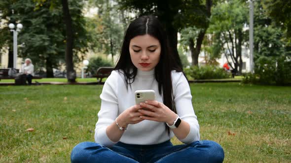 Young Woman Sitting on a Green Lawn in the Summer Park with a Smartphone
