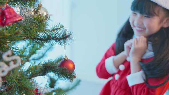 Asian young happy girl kid wear Santa hat and decorate Christmas tree in living room in house.