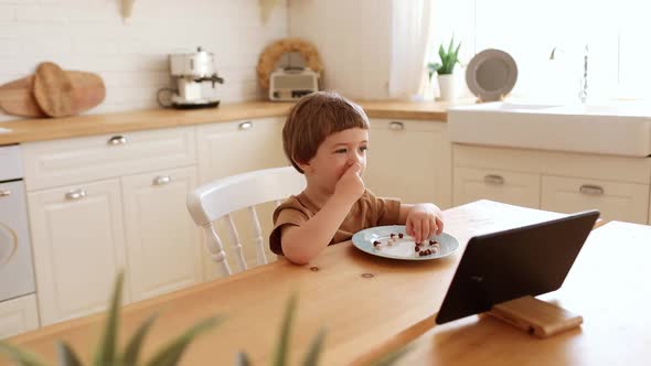 a Little Caucasian Boy Eats Krunchy and Watches Tablet PC at Kitchen Table