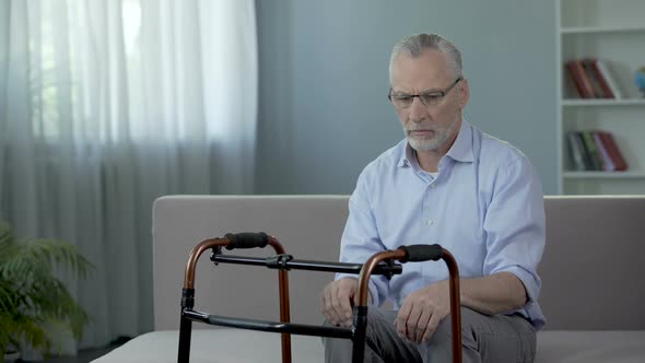 Old Male Sitting on Couch and Looking at Walking Frame Standing in Front of Him