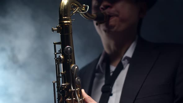 Saxophonist in the smoke plays a saxophone a dark studio. Male musician in suit and hat blows on sax