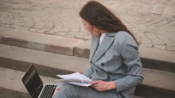 Young Happy Woman Sitting on Stairs While Using Laptop Working with Papers Documents on the Street