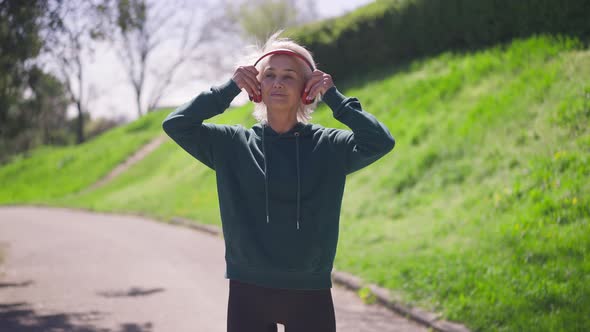 Senior Confident Sportswoman Putting on Headphones Putting Hands on Hips Smiling Looking at Camera