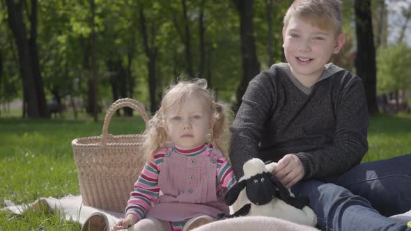 Portrait Teenage Blond Boy Sitting on Blanket in the Park Reading the Book To Her Little Sister in