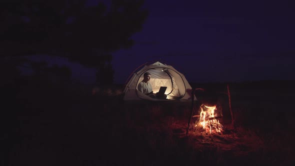 Freelancer Works Behind a Laptopin in a Tent Near a Fire