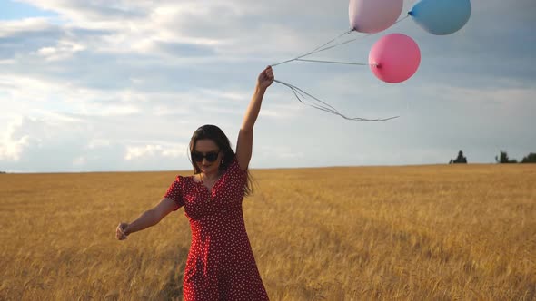 Happy Young Girl with Brown Hair Walking Through Golden Wheat Field and Holding Balloons in Hand