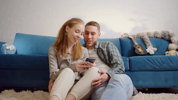 Couple Use Smartphone Device While Sitting on a Couch in the Cozy Apartment