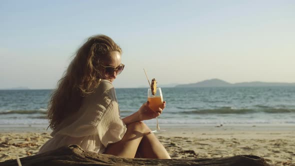 Woman Sitting in White Dress Drinks Pineapple Cocktail Pina Colada on Beach