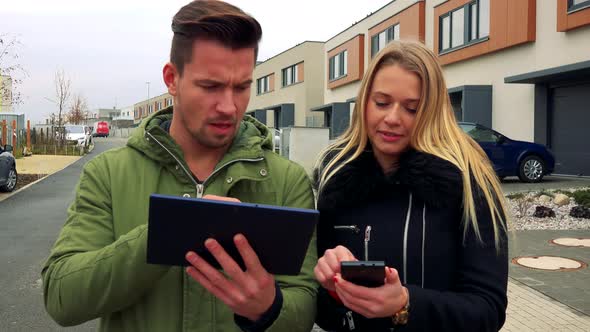 A Man and a Woman Discuss Something About a Tablet and a Smartphone on a Road in a Neighborhood