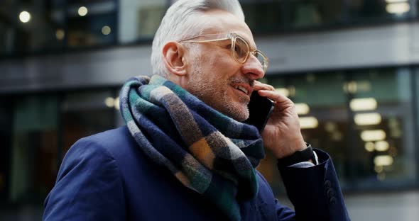 A Grayhaired Man Uses a Mobile Phone Standing in Business Center of the City