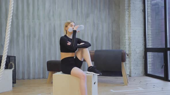 A Young Blonde Athletic Woman Drinks Water From A Bottle After An Exercise, Sits on a Jump Box