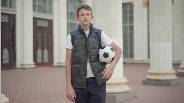 Portrait of Brunette Caucasian Schoolboy Posing with Soccer Ball in Front of School Building