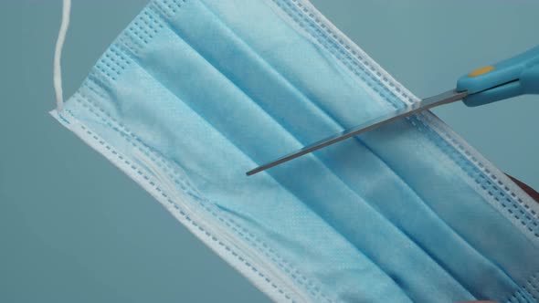 Scissors Cutting a Surgical Face Mask Against Coronavirus Disease on a Blue Background