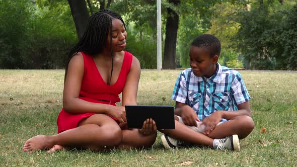 A Young Black Mother and Her Son Sit on Grass in a Park and Work on a Tablet