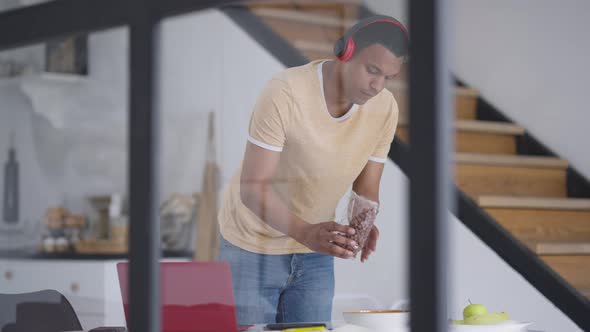 Young African American Man Pouring Cocoa Balls in Bowl Standing at Table in Home Office Kitchen