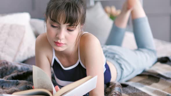 Medium Shot Focused Young Woman Reading Book Lying Crossing Legged on Bed at Home Interior