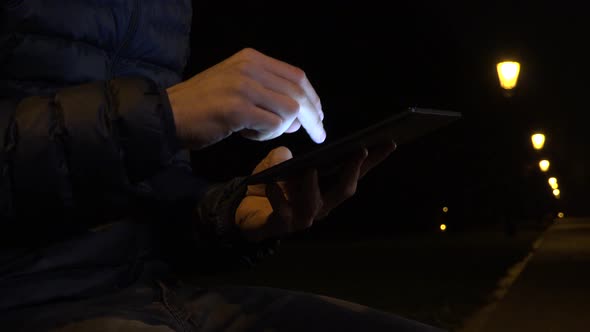 A Man Sits on a Bench and Works on a Tablet in an Urban Area at Night - Closeup - Streetlights