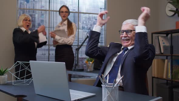 Cheerful Mature Old Businessman Raising Hands Celebrating Sudden Victory with Colleagues in Office