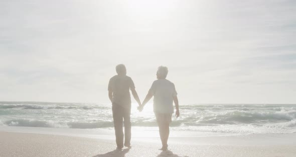Back view of hispanic senior couple holding hands, walking on beach at sunset