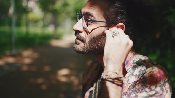 Serious curly-haired bearded man in eyeglasses putting on headphones on the bench