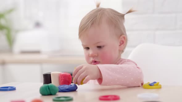 Cute Little Girl with Funny Ponytails on Head Sitting in Kitchen and Playing Multicolored Plasticine