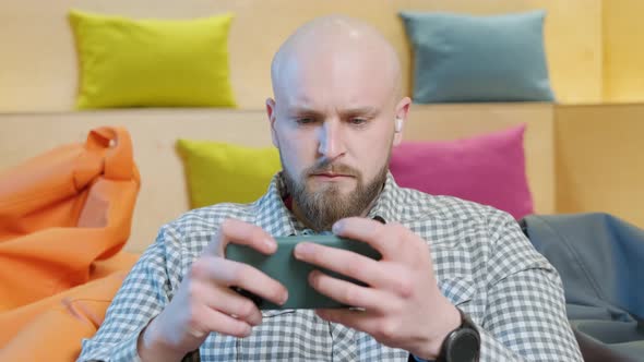 Close Up of an Young Man Sits on a Bean Bag While Playing Online Using His Phone