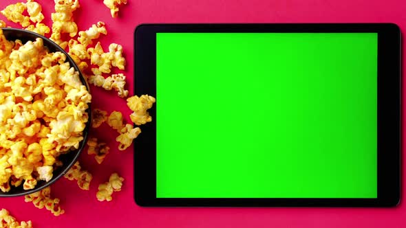 Tablet with green touch screen and popcorn in a bowl on red background.