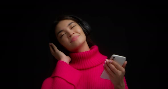 Woman in a Bright Pink Sweater Listens to Music with Headphones Through Phone