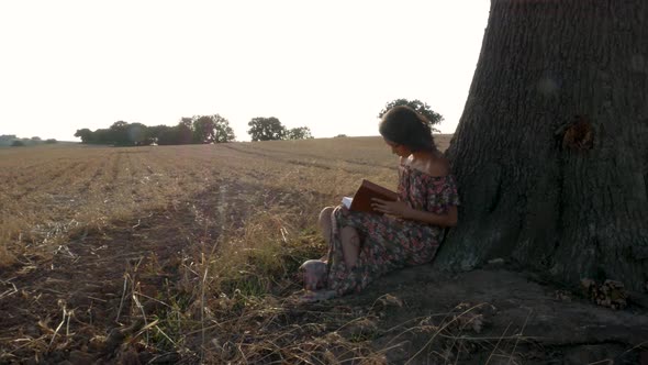 Tight shot of a young woman leaning up against a tree and reading in the setting sun.