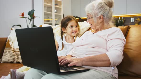 Grandmother is Smiling Typing on Laptop Talking to Granddaughter Sitting on Sofa in Studio Flat with