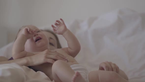 Close-up of a Beautiful Baby Girl Crying in Mother's Arms Laying in the Bed. A Woman Trying To Calm