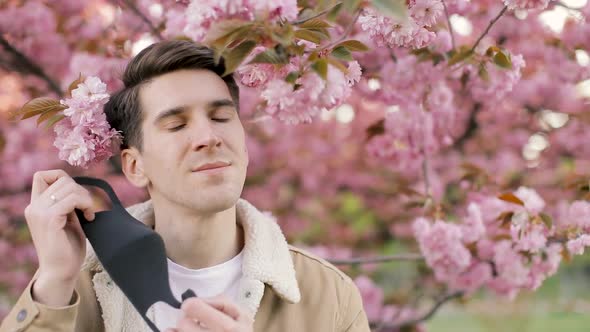 Man taking off protective face mask look at camera near sakura blossom. COVID-19, coronavirus