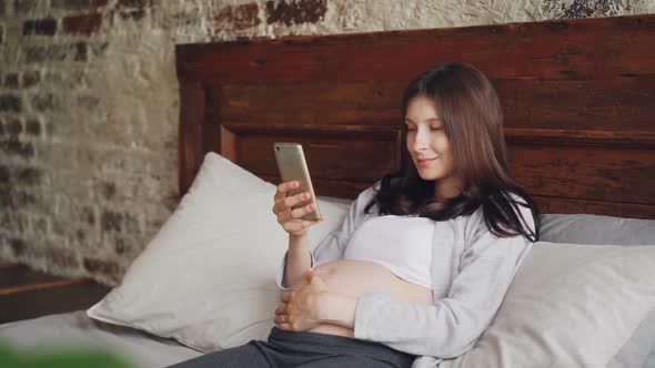 Attractive Young Woman Expecting Mother Is Using Smartphone, Stroking Her Belly and Smiling Lying in