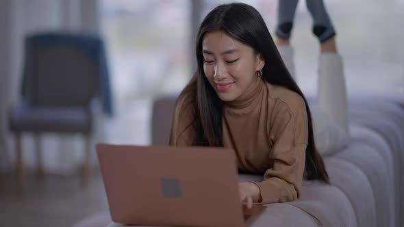 Smiling Asian Young Woman Lying on Couch Typing on Laptop Keyboard