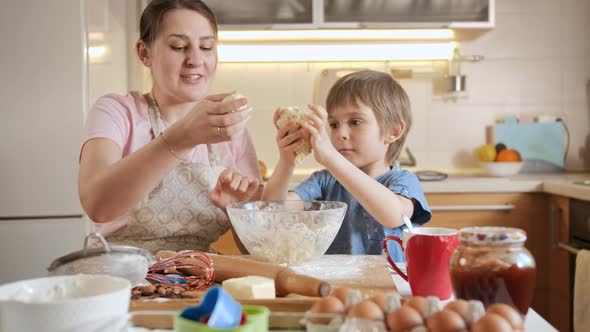 Young Mother with Little Son Kneading Dough and Forming It to Roll on Wooden Board