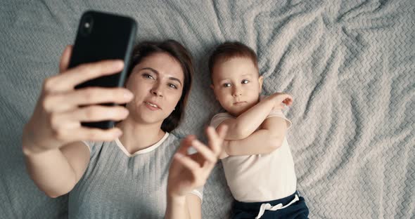 Mother and Her Baby Boy Making a Selfie or Video Call to Father or Relatives in a Bed