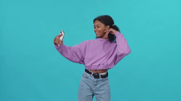 Cheerful African Girl Dances on a Blue Light Background and Takes a Selfie Video on Smartphone for a