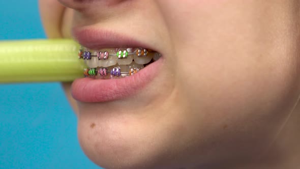 Teenage Girl with Braces on Her Teeth Eats Celery on a Blue Background. A Girl with Colored Braces