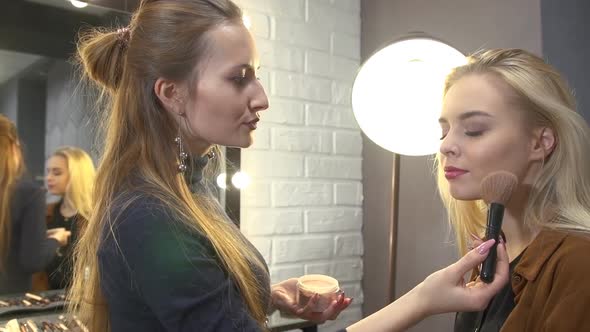 Young Stylist Putting Makeup on Face of Beautiful Blonde Girl