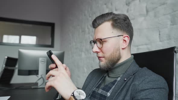 A Confident Young Man in Smart Casual Wear Is Holding Smart Phone looking at It While and Sitting