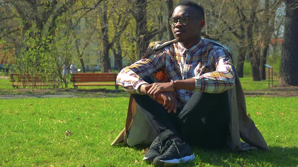 A Young Black Man in Glasses Sits on Grass in a Park on a Sunny Day and Looks Around